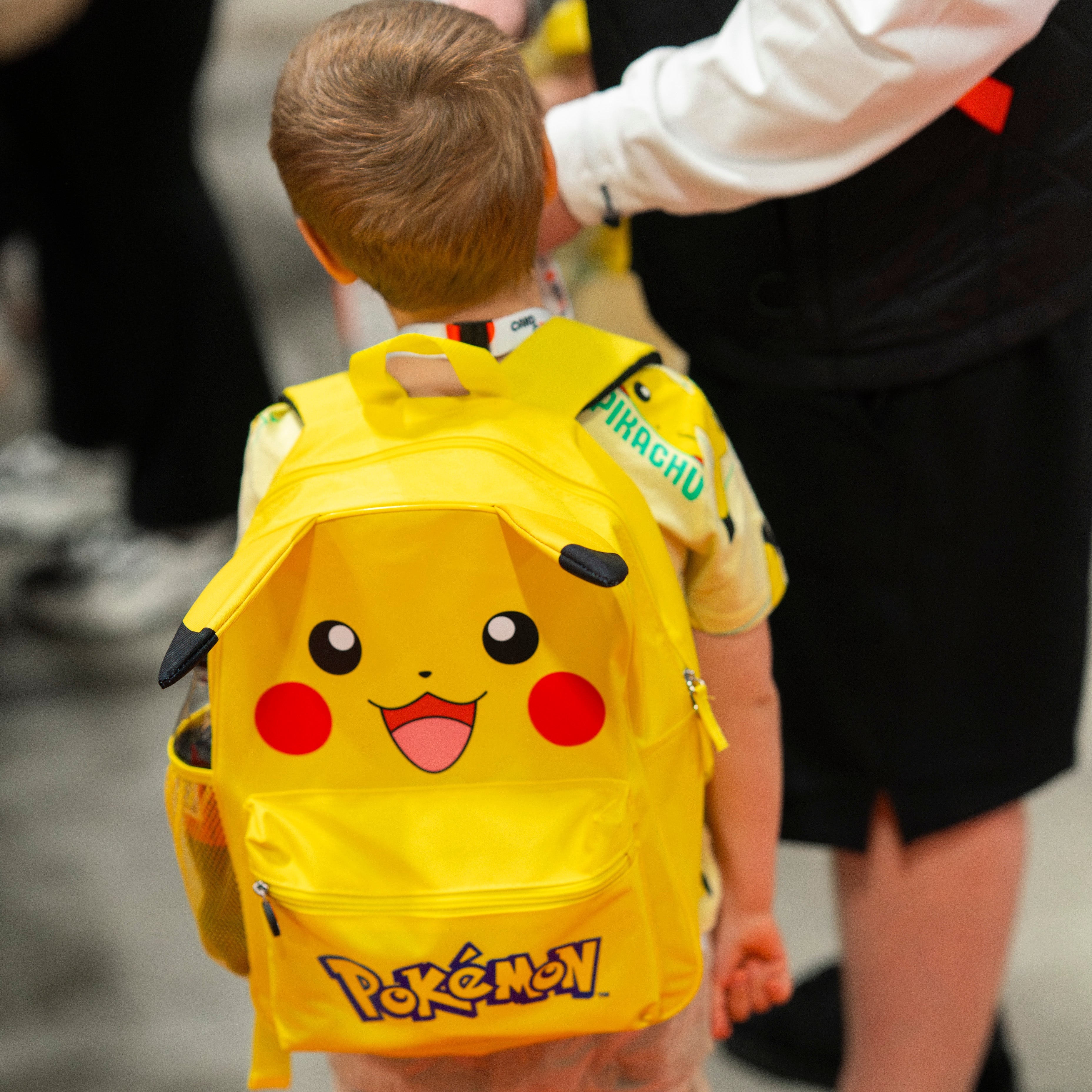 Child wearing a yellow Pokémon backpack with a Pikachu design, standing next to an adult.